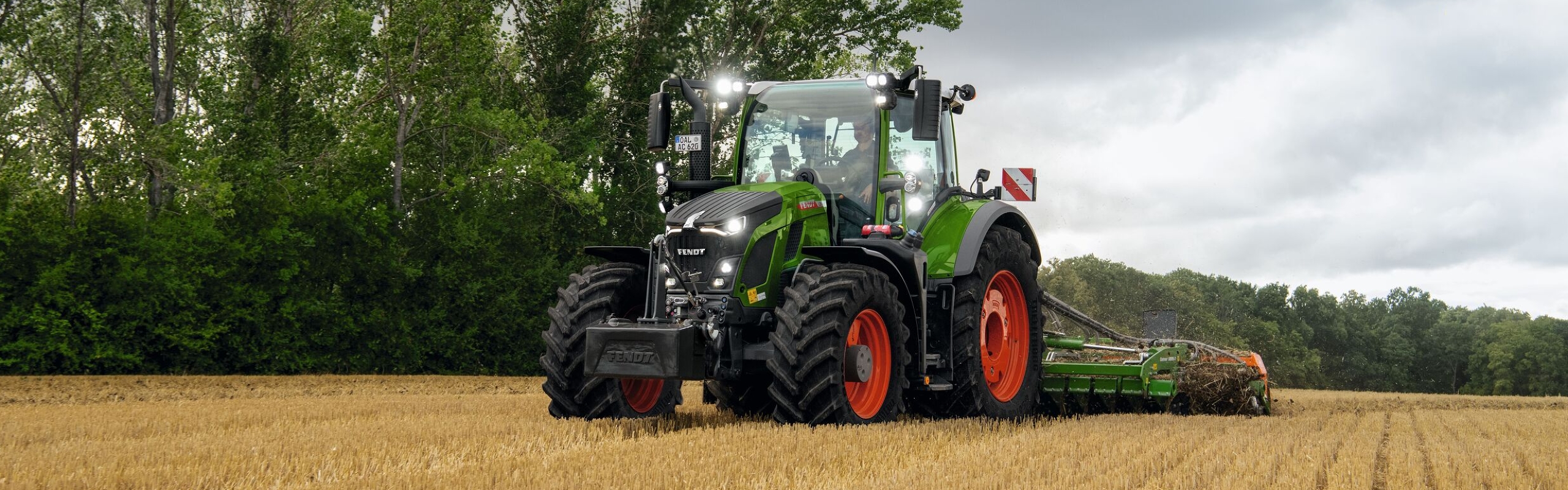 Il trattore Fendt 600 Vario verde con cerchi rossi in uso su un campo con alberi sullo sfondo e un cielo nuvoloso.