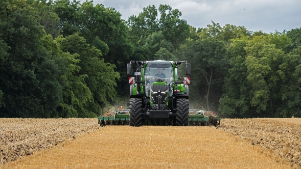 Il Fendt 600 Vario verde è in uso con un attrezzo in un campo marrone. Sullo sfondo una foresta verde scuro e un cielo nuvoloso.