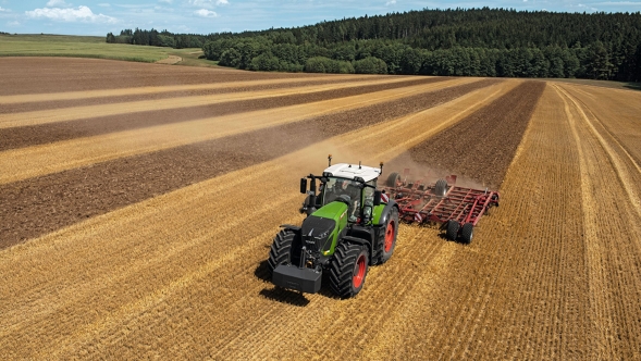 „Fendt 900 Vario“ „Horsch“ žemės įdirbimo įranga žemę įdirbantis žalias „Fendt 900 Vario“. Fone matosi medžiai ir mėlynas dangus.