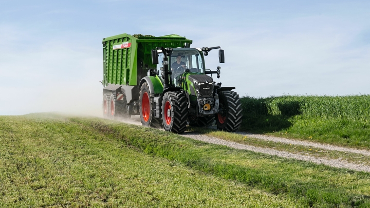 Nedideliu kaimo keliuku važiuoja žalias „Fendt 600 Vario“ traktorius raudonais ratlankiais ir žalia krautuvine priekaba „Fendt Tigo“, fone – priekyje ir užpakalyje – žalia pieva.
