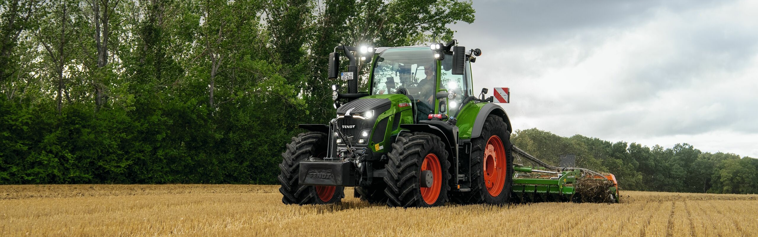 Lauke su medžiais (fone – debesuotas dangus) dirba žaliasis „Fendt 600 Vario“ traktorius raudonais ratlankiais.