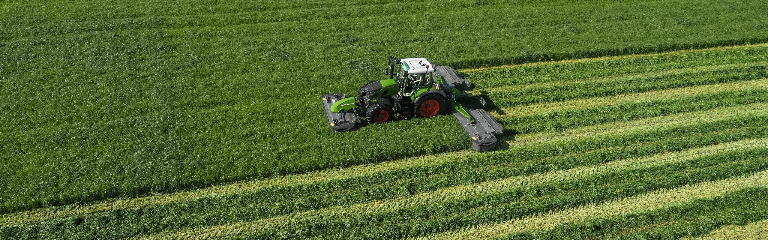 Dirbančio žaliojo traktoriaus „Fendt 600 Vario“ su žalia šienapjove „Fendt Slicer“ vaizdas iš paukščio skrydžio didelėje žalioje pievoje.