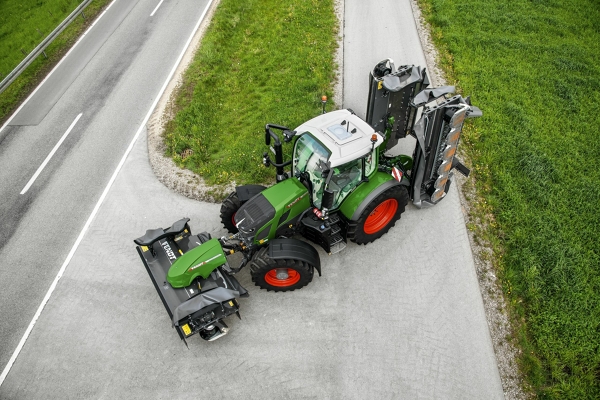 „Fendt 600 Vario“ Pilka gatve nedidele posūkio trajektorija važiuoja žalias „Fendt 600 Vario“ traktorius raudonais ratlankiais ir užlenktu padargu.