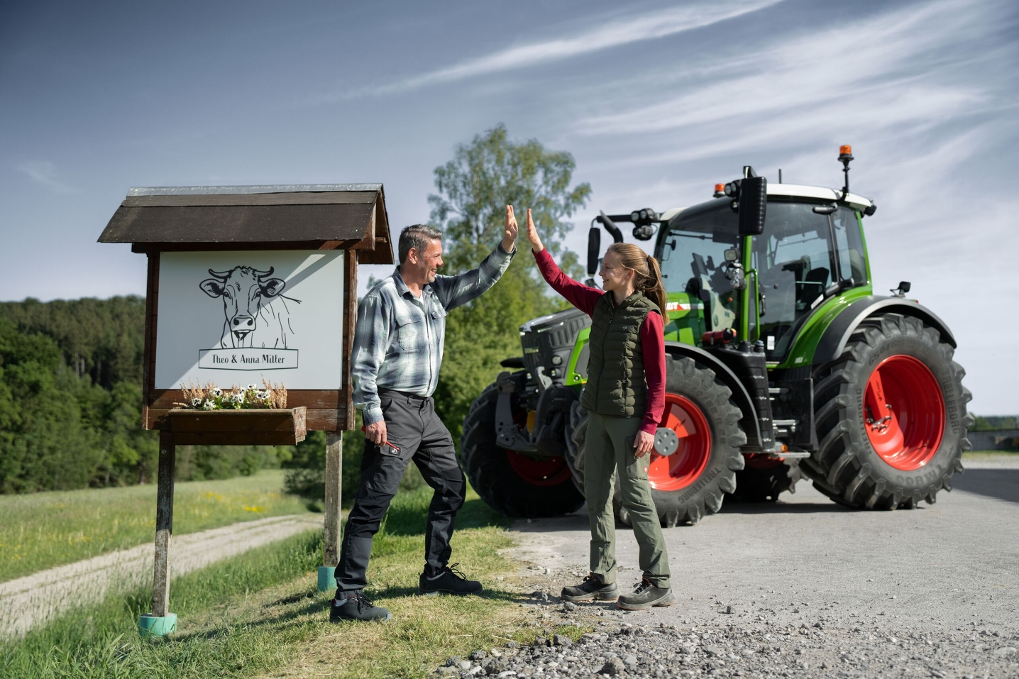 Ūkininkas ir jo dukra stovi priešais savo ūkio iškabą, fone jų „Fendt 500 Vario“