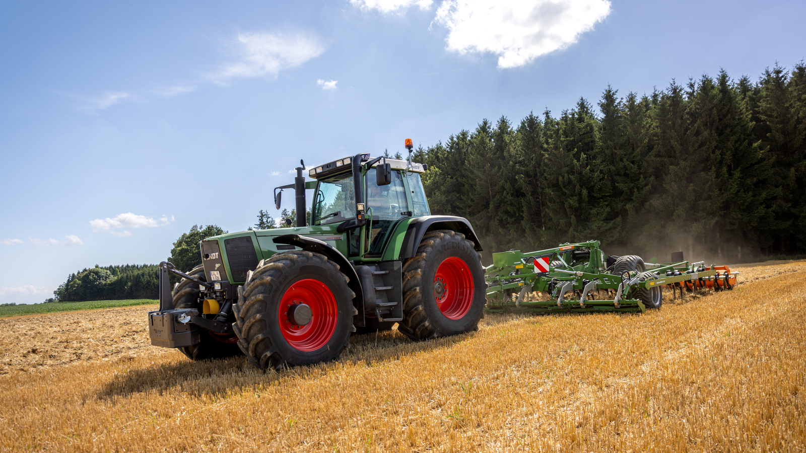 „Fendt Favorit 926 Vario“ Ūkininkas važiuoja per lauką su „Fendt Favorit 926 Vario“; fone matyti miškas.