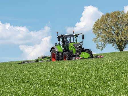Žalios spalvos „Fendt 700 Vario“ Gen7 šienauja pievą Fendt priekine ir galine žoliapjove; fone matyti medis.