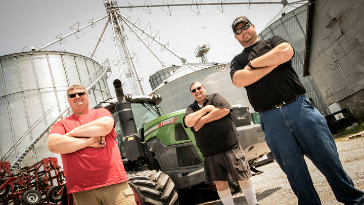Trīs lauksaimnieki stāv ar rokām krustā priekšā lielam Fendt traktoram un citām lauksaimniecības mašīnām.