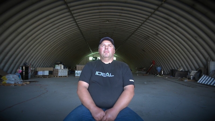 Lielā zālē stāvoša lauksaimnieka portrets, kurš valkā melnu T-kreklu ar uzrakstu IDEAL.