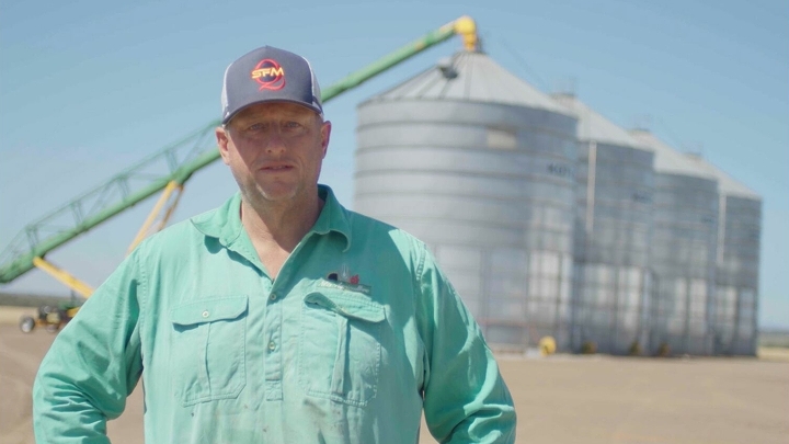 Marka Beikera portrets, kurā viņš redzams savā saimniecībā, tērpies gaiši zilā kreklā un cepurē, fonā redzams lauksaimniecības tehnikas un zils debess.