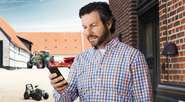 Een man staat op een erf met Fendt-tractoren op de achtergrond en kijkt naar zijn smartphone, die hij in zijn hand houdt.