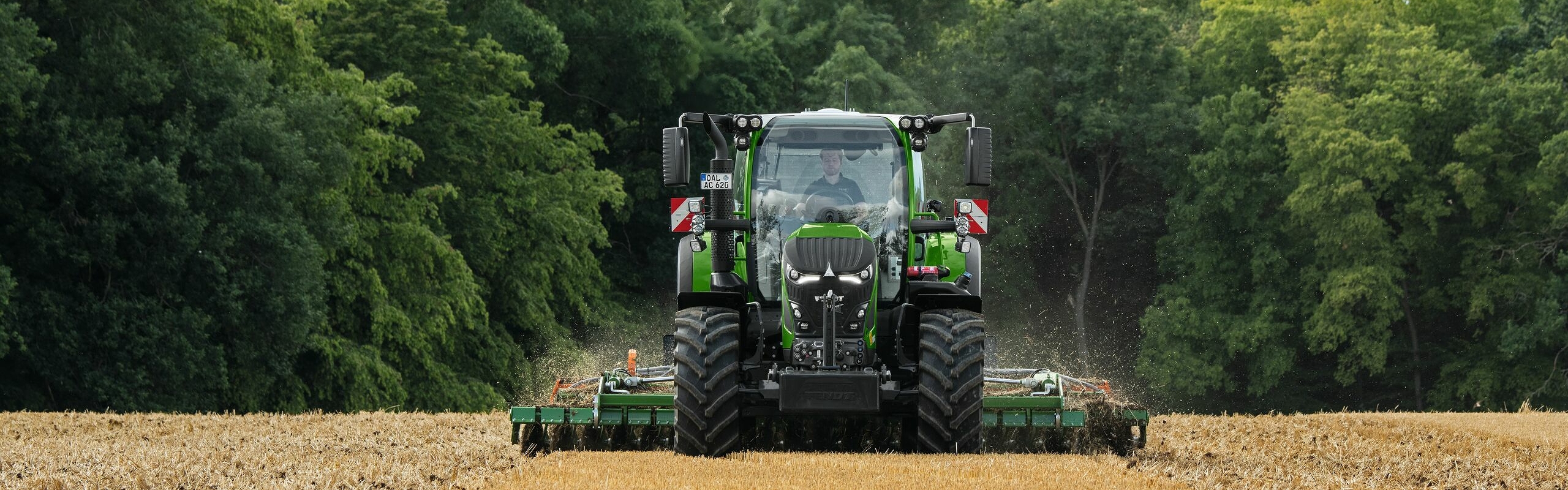 De groene Fendt 600 Vario met aanbouwapparatuur aan het werk op een bruin veld. Op de achtergrond is een donkergroen bos en een bewolkte lucht te zien.