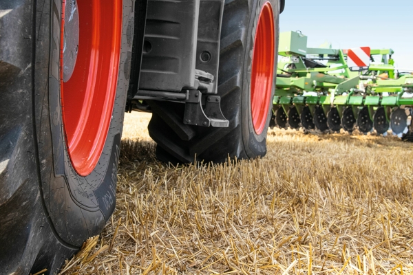 Tractorbanden Close-up van twee zwarte tractorbanden met rode velgen, die op een lichtbruin veld staan. Op de achtergrond ziet men groene Fendt aanbouwapparatuur.