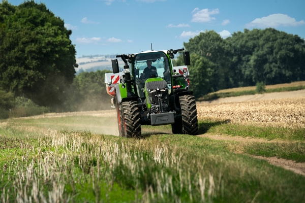 De groene Fendt 600 Vario-trekker met rode velgen rijdt op een kleine veldweg met een groene weide op de voorgrond en een donkergroen bos en een lichtblauwe lucht op de achtergrond.