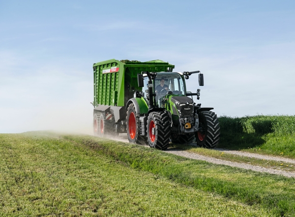 Der grüne Fendt 600 Vario Traktor mit roten Felgen und dem grünen Ladewagen Fendt Tigo fährt auf einer kleinen Feldstraße mit einer grünen Wiese im Vorder- und Hintergrund.