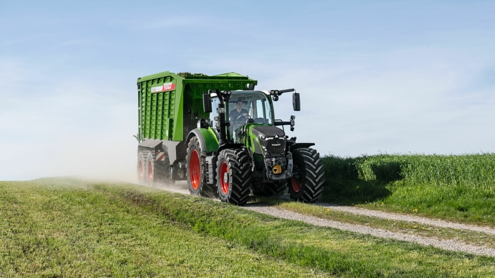 De groene tractor Fendt 600 Vario-trekker met rode velgen en de groen opraapwagen Fendt Tigo rijdt op een kleine veldweg met een groene weide op de voor- en achtergrond.