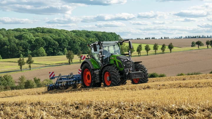 Een groene Fendt-tractor met rode velgen tijdens het bewerken van de grond op een geoogst veld met een zacht glooiend heuevellandschap op de achtergrond.