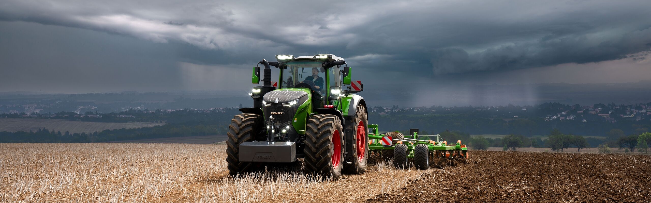 Onder een dreigende hemel met onweerswolken in de verte gaat een tractor, een Fendt 1000 Vario, onverstoorbaar verder met het werk op het veld