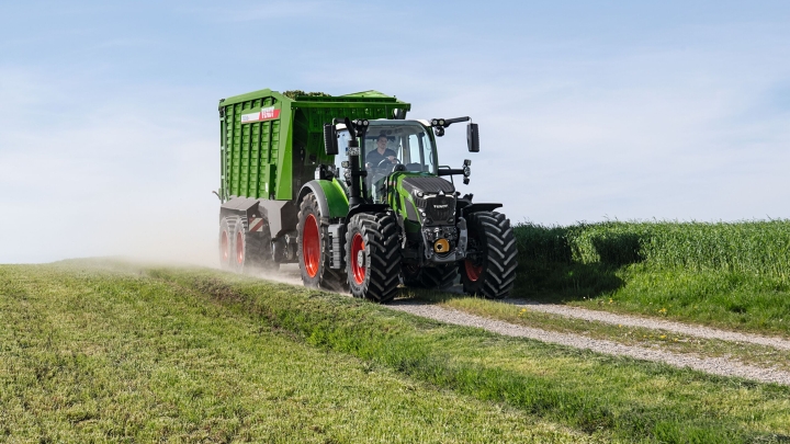De groene tractor Fendt 600 Vario-trekker met rode velgen en de groen opraapwagen Fendt Tigo rijdt op een kleine veldweg met een groene weide op de voor- en achtergrond.