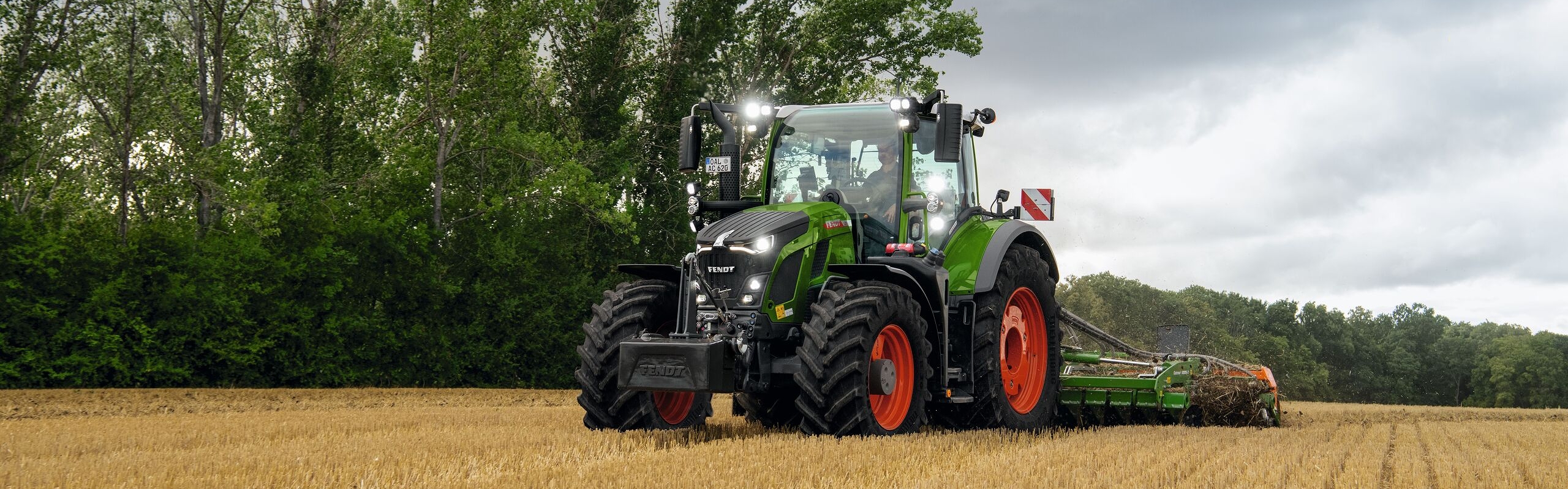 De groene Fendt 600 Vario met rode velgen aan het werk op een akker met bomen en een bewolkte lucht op de achtergrond.