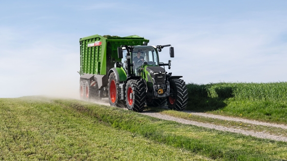De groene tractor Fendt 600 Vario-trekker met rode velgen en de groen opraapwagen Fendt Tigo rijdt op een kleine veldweg met een groene weide op de voor- en achtergrond.