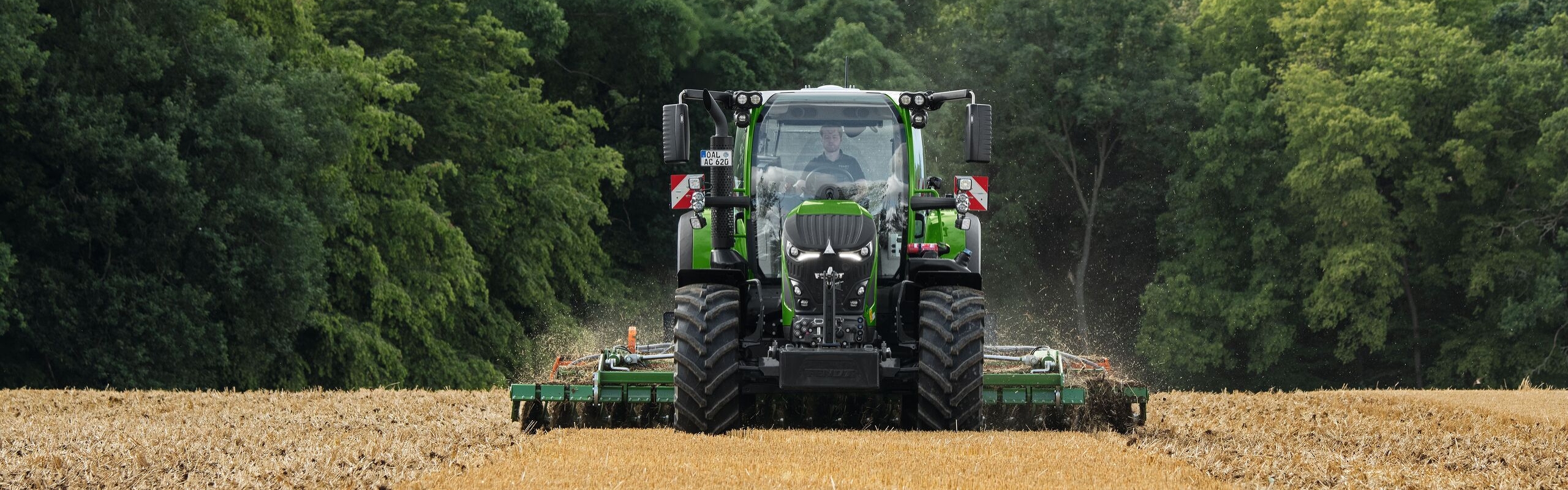 De groene Fendt 600 Vario met aanbouwapparatuur aan het werk op een bruin veld. Op de achtergrond is een donkergroen bos en een bewolkte lucht te zien.