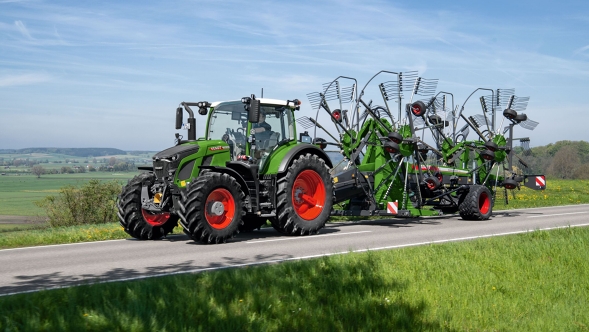 De groene tractor Fendt 600 Vario met rode velgen rijdt met de groene Fendt Former op een grijze weg. Op de achtergrond is een grote groene weide en een blauwe lucht te zien.