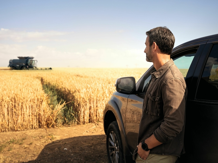 Een boer staat tegen een auto geleund voor zijn graanveld en kijkt tevreden toe, hoe een Fendt-maaidorser oogst