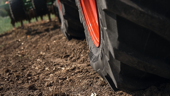Close-up van een zwaar doorgezakte grote tractorband met zwart profiel en rode velg op los bruin akkerland. Op de achtergrond is onscherp een agrarisch aanbouwerktuigw te herkennen.