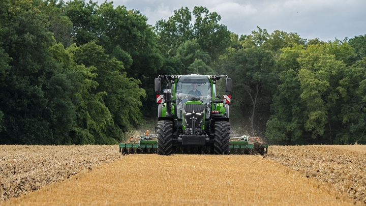 De groene Fendt 600 Vario met aanbouwapparatuur aan het werk op een bruin veld. Op de achtergrond is een donkergroen bos en een bewolkte lucht te zien.