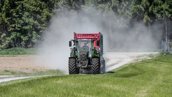 Een Fendt 724 Vario Gen7.1 vervoert een Strautmann opraapwagen op de veldbaan. Een groene Fendt 724 Vario Gen7.1 in combinatie met een Strautmann opraapwagen rijdt over een zandpad recht op de camera af. Op de achtergrond is een bos te zien.
