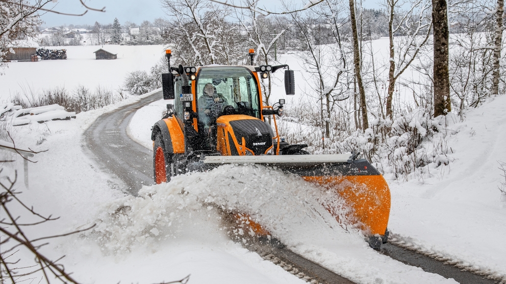 Orange Fendt-traktor rydder snø på en vei i et snølandskap