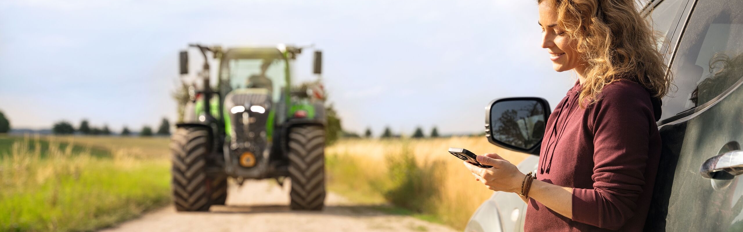 En kvinne med lyse krøller og mørkerød genser lener seg mot en bil og ser på mobiltelefonen sin. I bakgrunnen ser vi en Fendt-traktor.