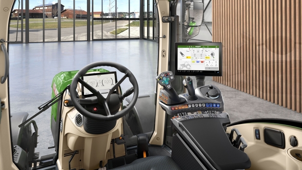 Innsiden av VisioPlus-førerhuset på en Fendt 300 Vario Gen5 med ergonomisk kjørespak, ratt og stor terminal foran en moderne gård med glassfasade