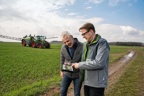 Dwóch rolników sprawdza dane ciągnika Fendt Rogator 300, który stoi za nimi, na iPadzie w FendtONE.