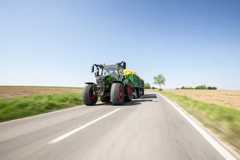 Zielony Fendt 500 Vario jedzie drogą z dwiema zielonymi przyczepami z żółtą plandeką.