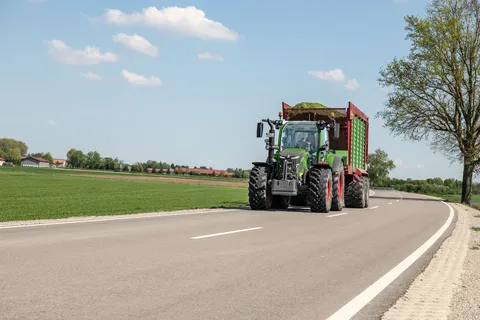 Zielony Fendt 500 Vario z pełną przyczepą zbierającą przejeżdża po ulicy w kierunku kamery.