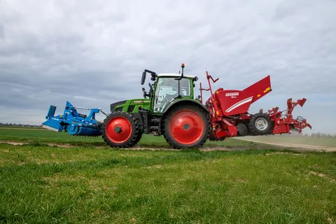 Zielony Fendt 500 Vario z czerwoną sadzarką do ziemniaków z tyłu i niebieską broną talerzową z przodu jedzie po polnej drodze.