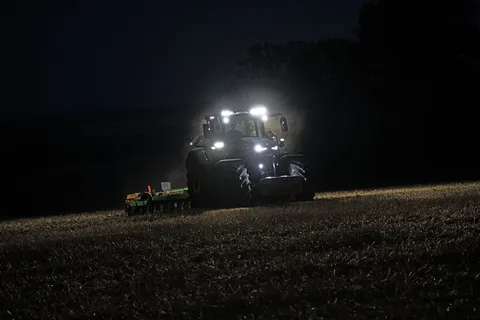 Ciągnik Fendt 800 jedzie po polu w ciemności z włączonymi reflektorami, ciągnąc bronę talerzową.