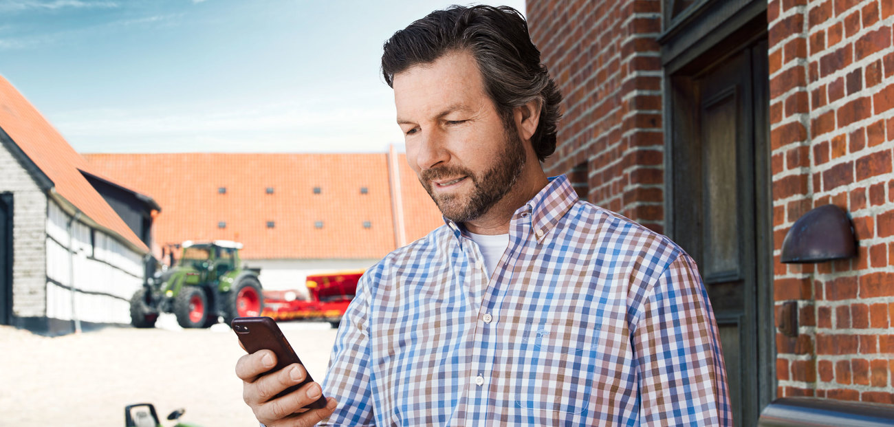 Mężczyzna stoi na podwórku, z ciągnikami Fendt w tle, i patrzy na swój smartfon, który trzyma w ręku.