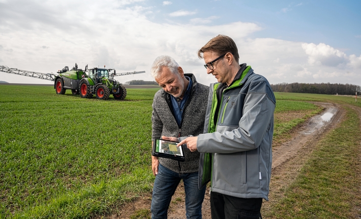 Dwóch rolników sprawdza dane ciągnika Fendt Rogator 300, który stoi za nimi, na iPadzie w FendtONE.