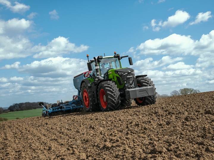 Zielony ciągnik Fendt 900 Vario z czerwonymi obręczami kół jedzie po brązowym polu, w tle niebieskie niebo z białymi chmurami.
