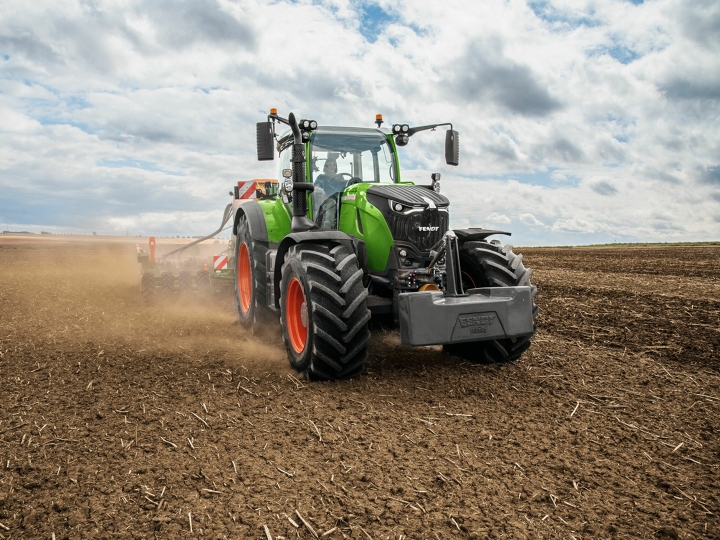 Zielony ciągnik Fendt 700 Vario z czerwonymi obręczami kół jedzie po brązowym polu, w tle niebieskie niebo z białymi chmurami.