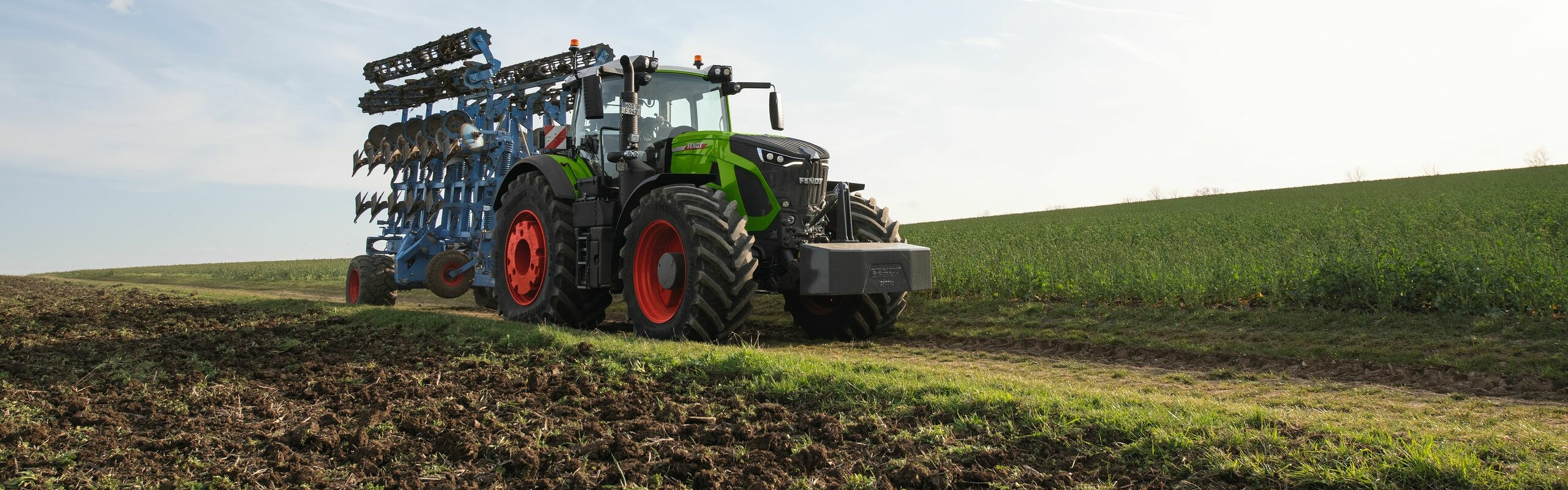Zielony ciągnik Fendt 900 Vario z czerwonymi obręczami kół i niebieskim elementem roboczym jedzie wąską polną drogą. W tle widać małe zielone wzgórze porośnięte trawą i jasnobłękitne niebo.