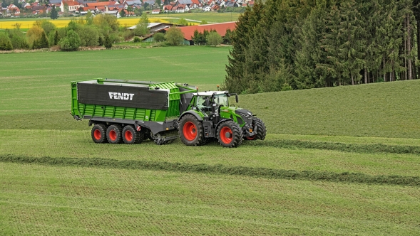 Zielony ciągnik Fendt 900 Vario z przyczepą zbierającą Fendt Tigo jedzie po zielonym polu. W tle widać ciemnozielony las i małą wieś.