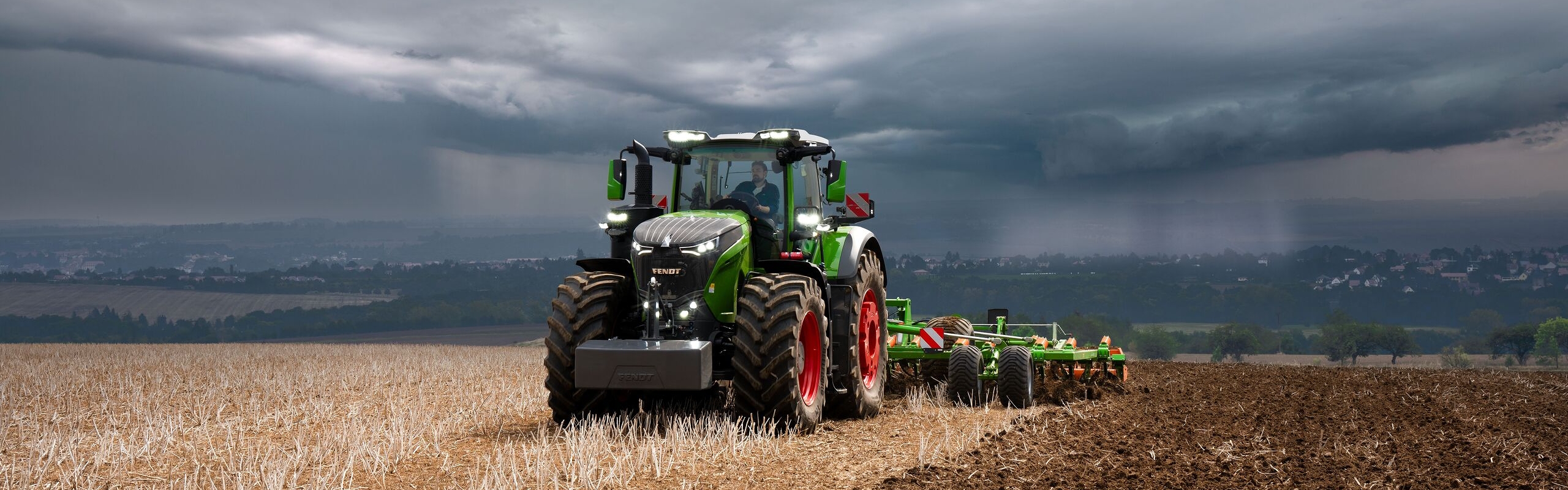 Przed nadciągającymi ciężkimi chmurami burzowymi z oddali ciągnik Fendt 1000 Vario wykonuje prace w polu nadal z pełną mocą