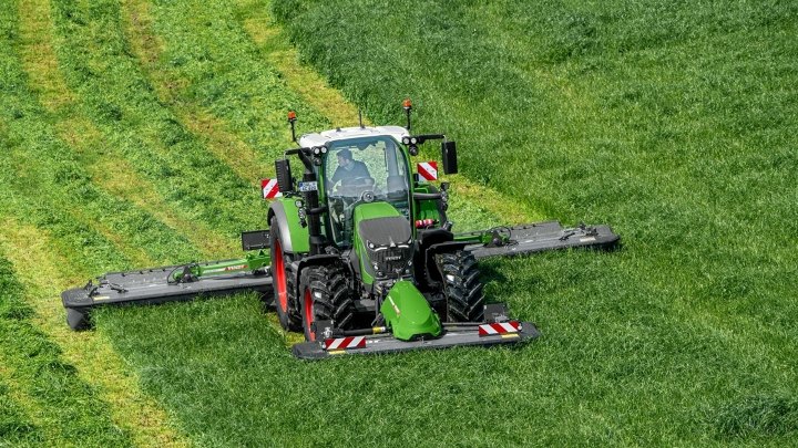 Zielony ciągnik Fendt 600 Vario ciągnik kosi zieloną łąkę z doczepioną zieloną kosiarką Fendt Slicer. W tle widać ślady koszenia.