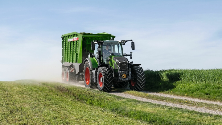 Zielony ciągnik Fendt 600 Vario z czerwonymi obręczami kół jedzie z zieloną przyczepą zbierającą Fendt Tigo małą polną drogą. Na pierwszym planie i w tle widać zieloną łąkę.