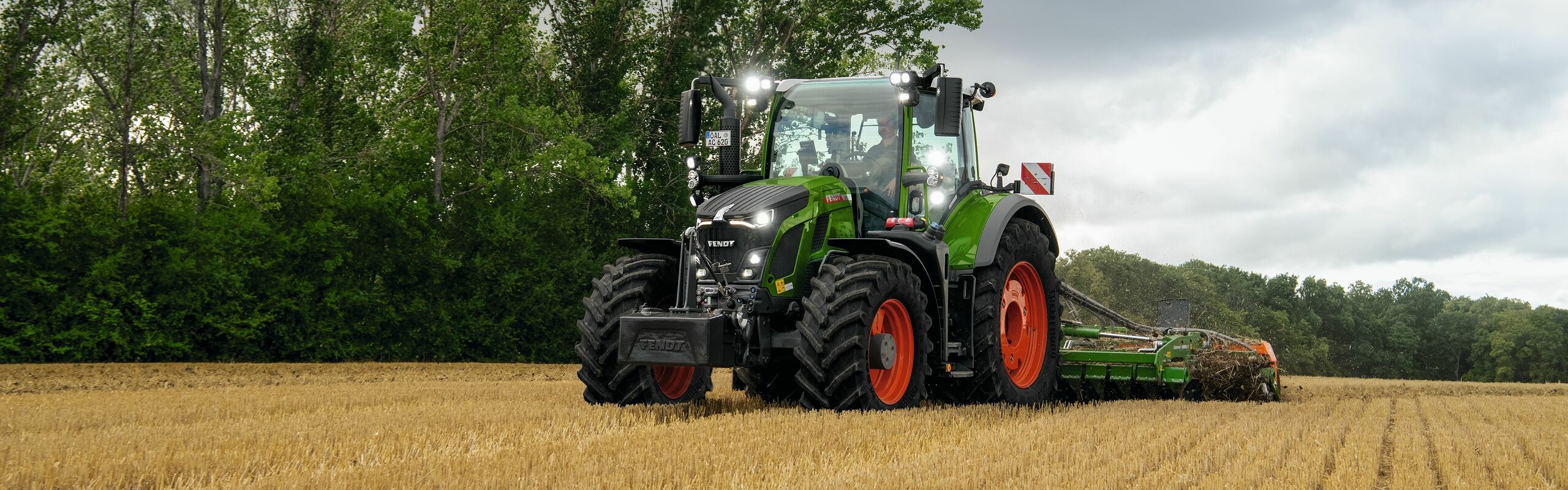 Zielony ciągnik Fendt 600 Vario z czerwonymi obręczami kół pracuje na polu. W tle drzewa i pokryte chmurami niebo.