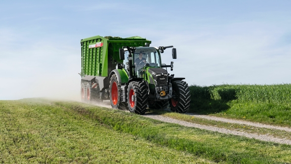 Zielony ciągnik Fendt 600 Vario z czerwonymi obręczami kół jedzie z zieloną przyczepą zbierającą Fendt Tigo małą polną drogą. Na pierwszym planie i w tle widać zieloną łąkę.