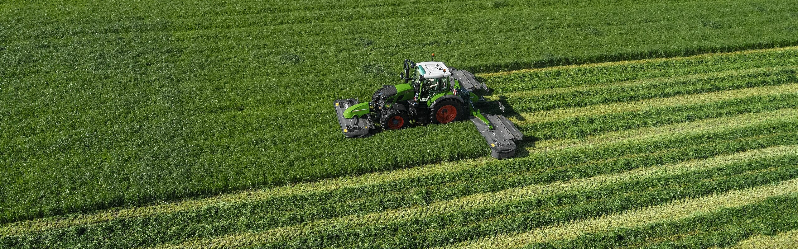 Widok z lotu ptaka na zielony ciągnik Fendt 600 Vario podczas pracy z zieloną kosiarką Fendt Slicer na dużej, zielonej łące.